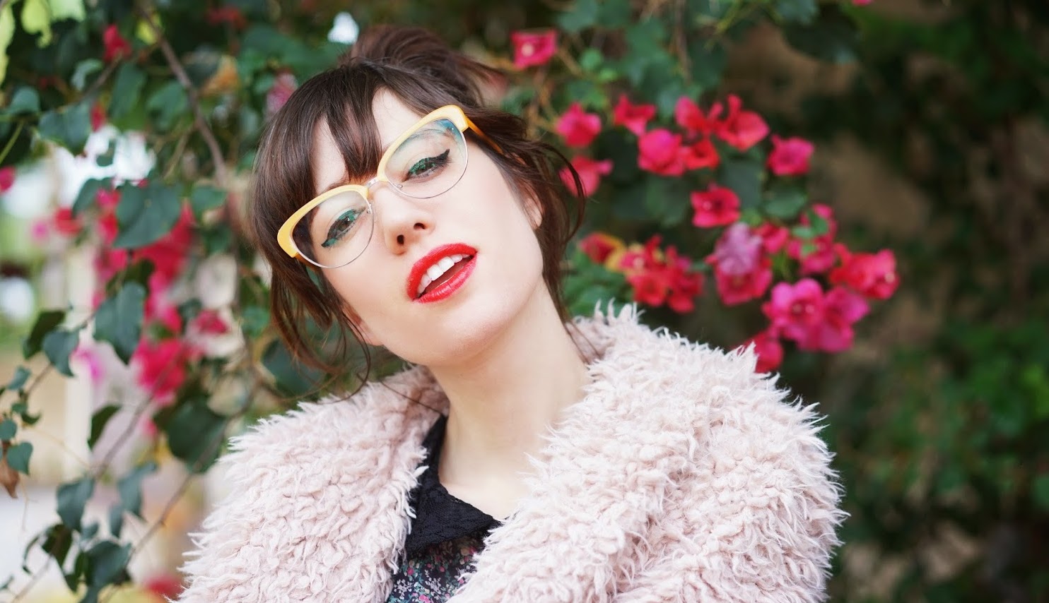 Woman wearing yellow-framed glasses and a fluffy beige coat, with red lipstick and floral background.