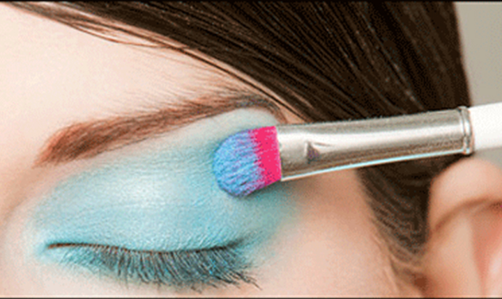 Applying blue eyeshadow with a brush featuring pink bristles on a closed eyelid.