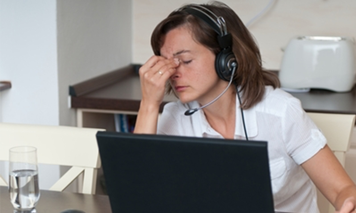 Woman wearing a headset, sitting at a desk with a laptop, looking stressed and pinching her nose.