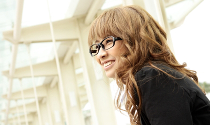 Woman with long, wavy hair wearing black rimmed glasses and a black jacket, smiling in a bright environment.