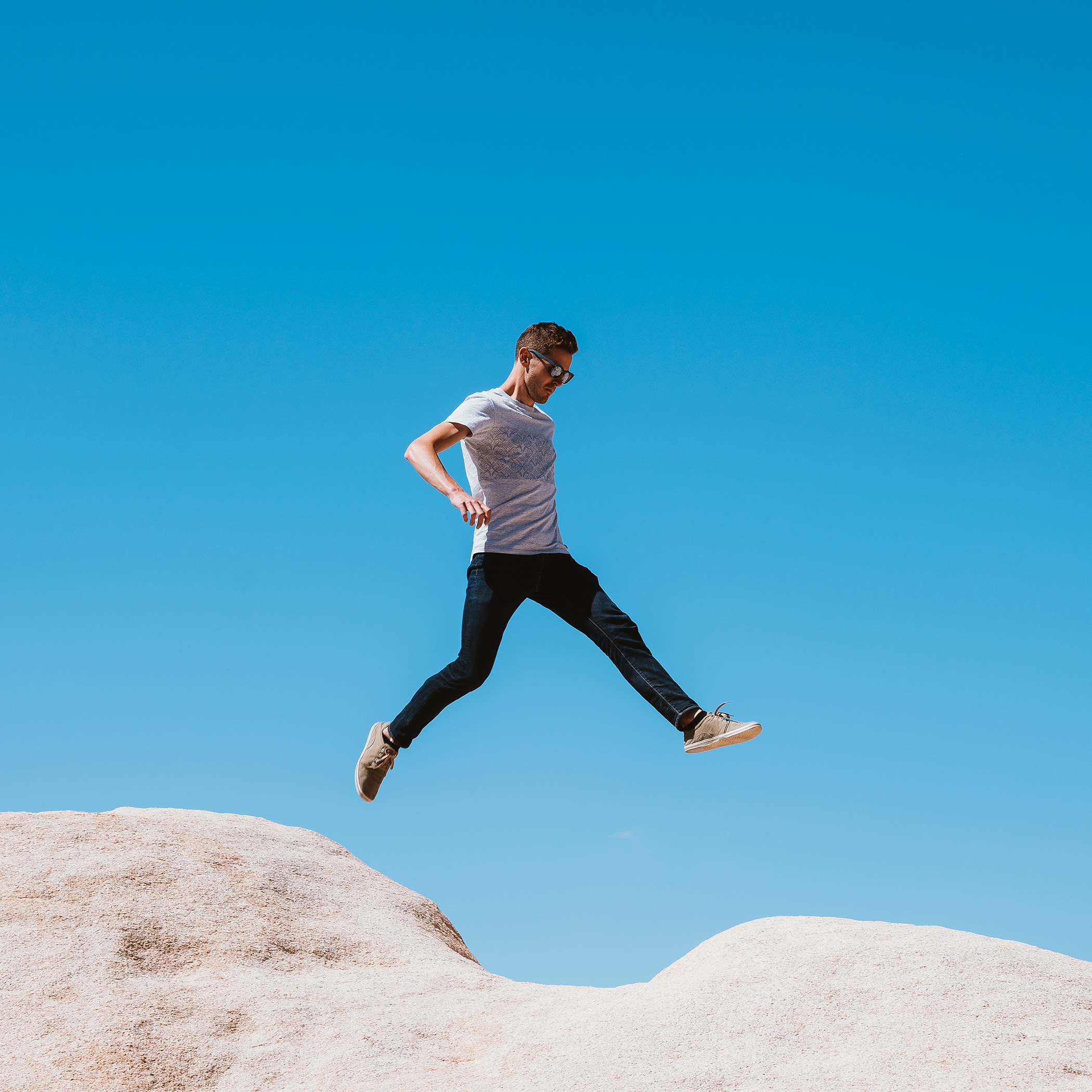 Man in casual attire mid-air leaping between two large rocks against a clear blue sky.