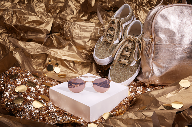 Gold glitter sneakers, gold backpack, pink sunglasses on a white block, and gold sequin earrings on gold background.