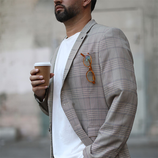 Man wearing a plaid blazer with eyeglasses in the chest pocket, holding a coffee cup.