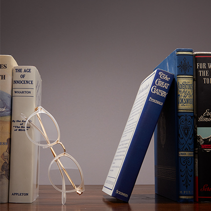 Clear-framed eyeglasses propped against a row of books. Titles include The Age of Innocence and The Great Gatsby.