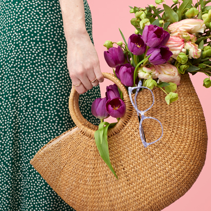 Woven straw handbag with round handles, filled with purple and pink tulips, and glasses hanging from it.