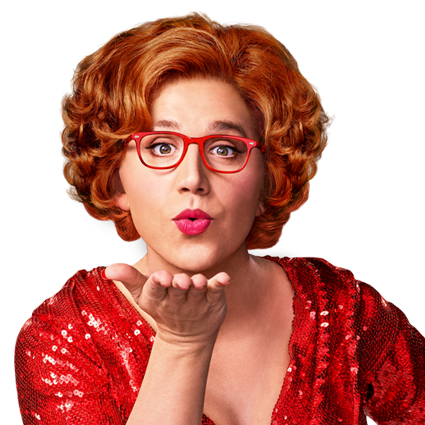 Person with red curly hair, wearing red glasses and a sequined red outfit, blowing a kiss.