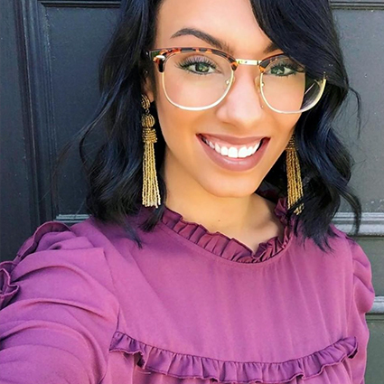 Woman wearing oversized, retro-style, gold-rimmed eyeglasses and long, gold tassel earrings.