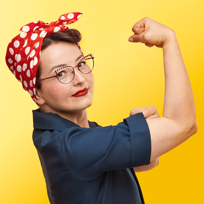 Person flexing arm wearing a red polka-dot headscarf, glasses, blue shirt, against yellow background.