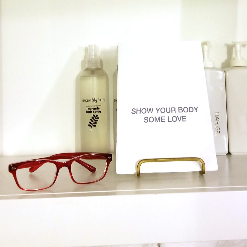 Red glasses, a bottle labeled "ginger lily farm miracle hair spray," and a sign saying "SHOW YOUR BODY SOME LOVE."