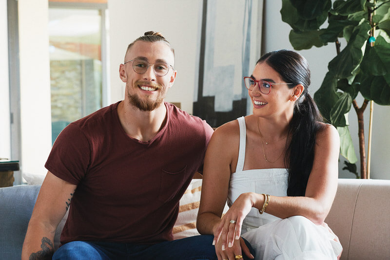 Two people wearing stylish eyeglasses sitting on a couch, both smiling and looking content.