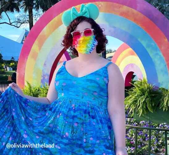 Person wearing a blue dress, red sunglasses, green mouse ears, and rainbow face mask. @oliviawiththeland