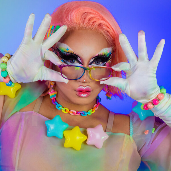 Person wearing colorful star accessories, white gloves, holding glasses with playful expression and vibrant makeup.