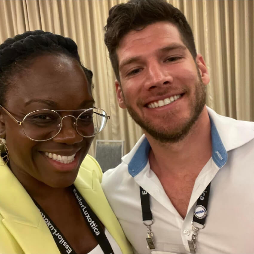 Two people smiling, wearing lanyards with text "EssilorLuxottica" and "Ele".