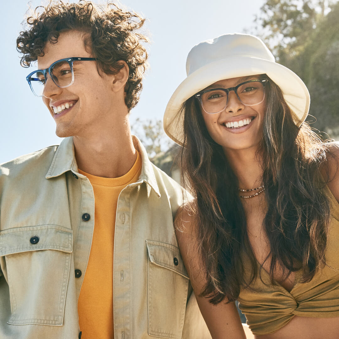 Man wearing blue rectangular eyeglasses, yellow shirt, and beige jacket; woman wearing beige bucket hat, tortoise-shell eyeglasses, and olive top.