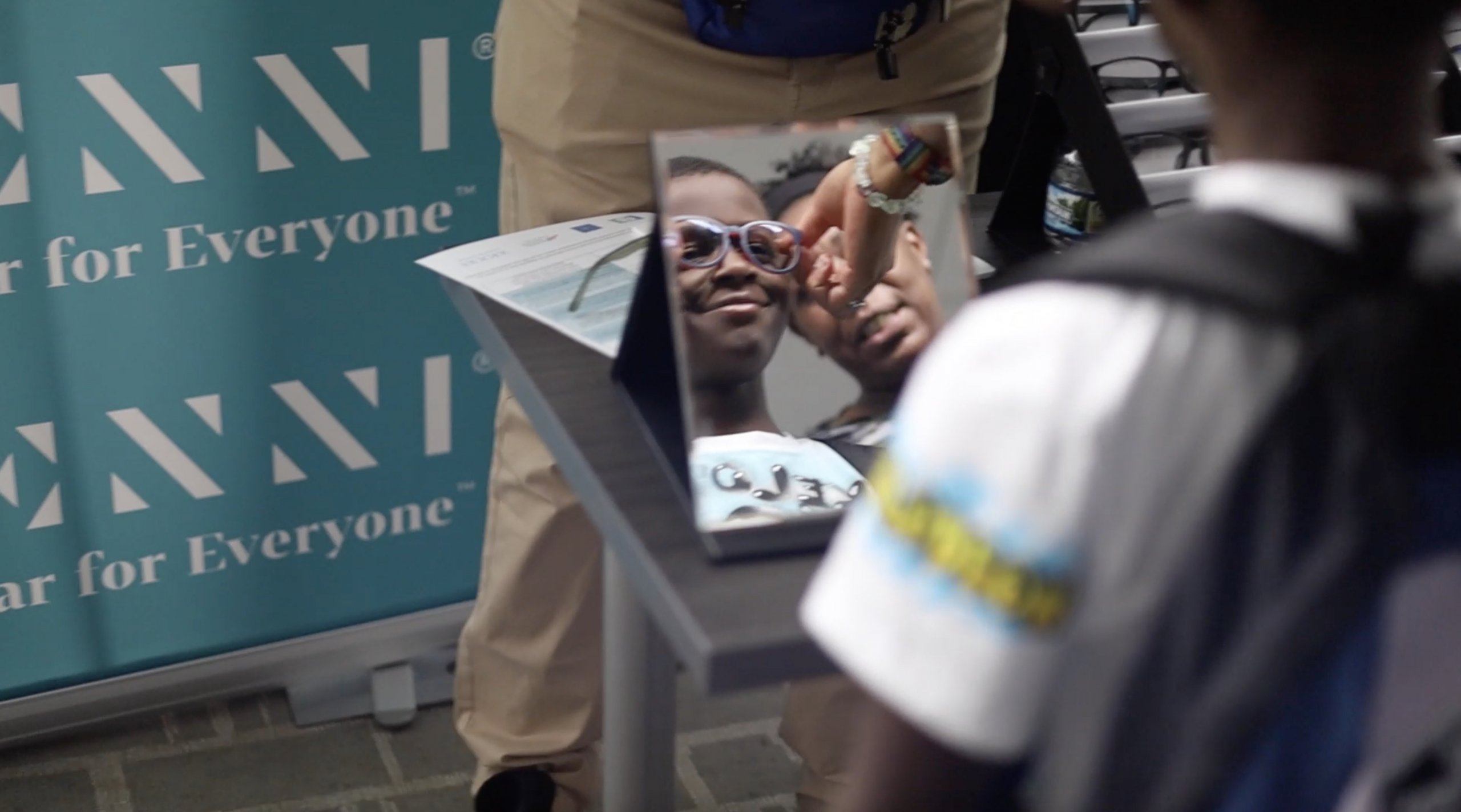 A child wearing glasses smiles at their reflection in a mirror by the ENNNI "Eyewear for Everyone" banner.