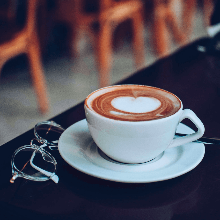 Latte in a white cup and saucer with a heart-shaped foam design, next to eyeglasses on a table.