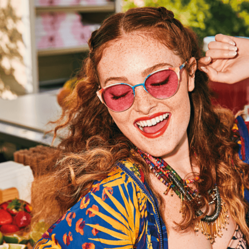 Woman wearing pink sunglasses, a colorful shirt, and layered necklaces, smiling and dancing.