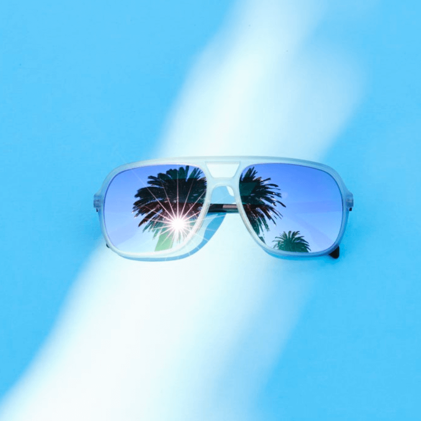 White-framed sunglasses with mirrored lenses reflecting palm trees and a bright sunburst against a blue background.