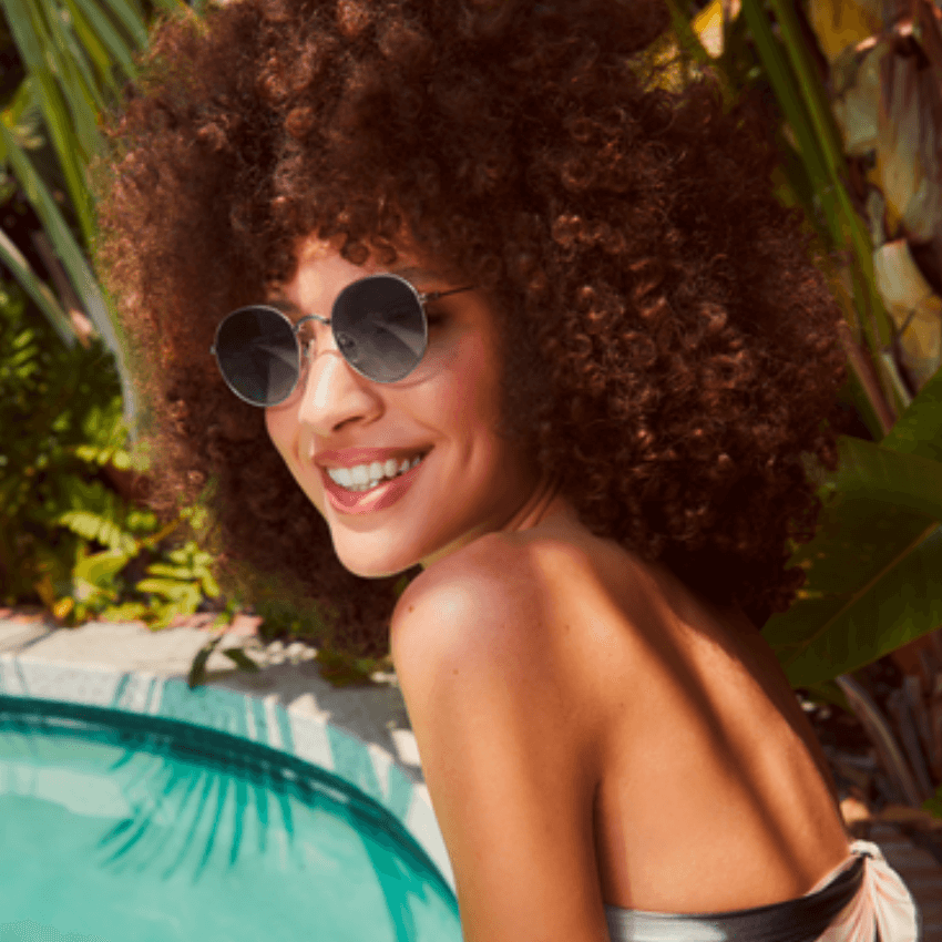 Woman wearing round sunglasses and smiling, with curly hair, next to a pool.