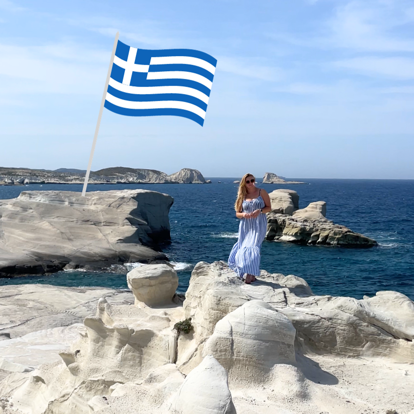 Woman in a striped dress standing on white rock formations by the sea, Greek flag in the background.