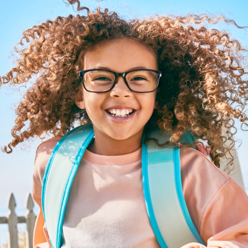 Smiling child with curly hair wearing glasses and a light blue backpack.