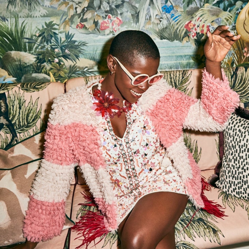 Woman in a pink and white textured sweater, floral necklace, sequined dress, and white sunglasses.