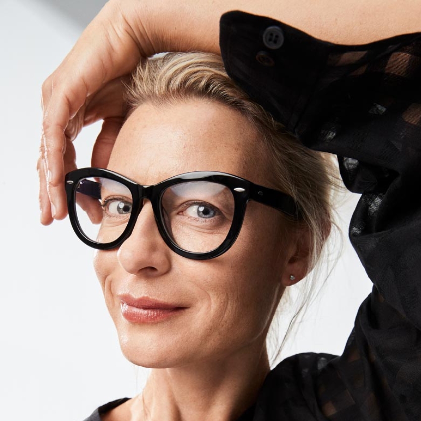 Woman wearing black-rimmed eyeglasses, lightly touching her forehead with her left hand.