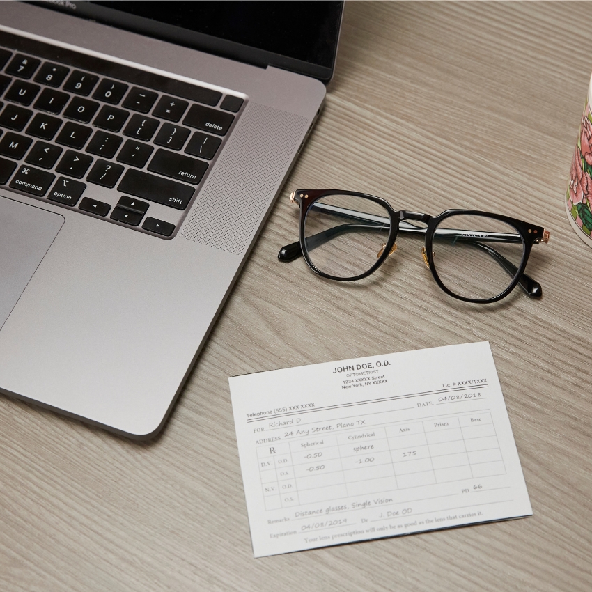 Prescription eyeglasses next to a laptop and an eyeglass prescription card from "JOHN DOE, O.D. OPTOMETRIST."