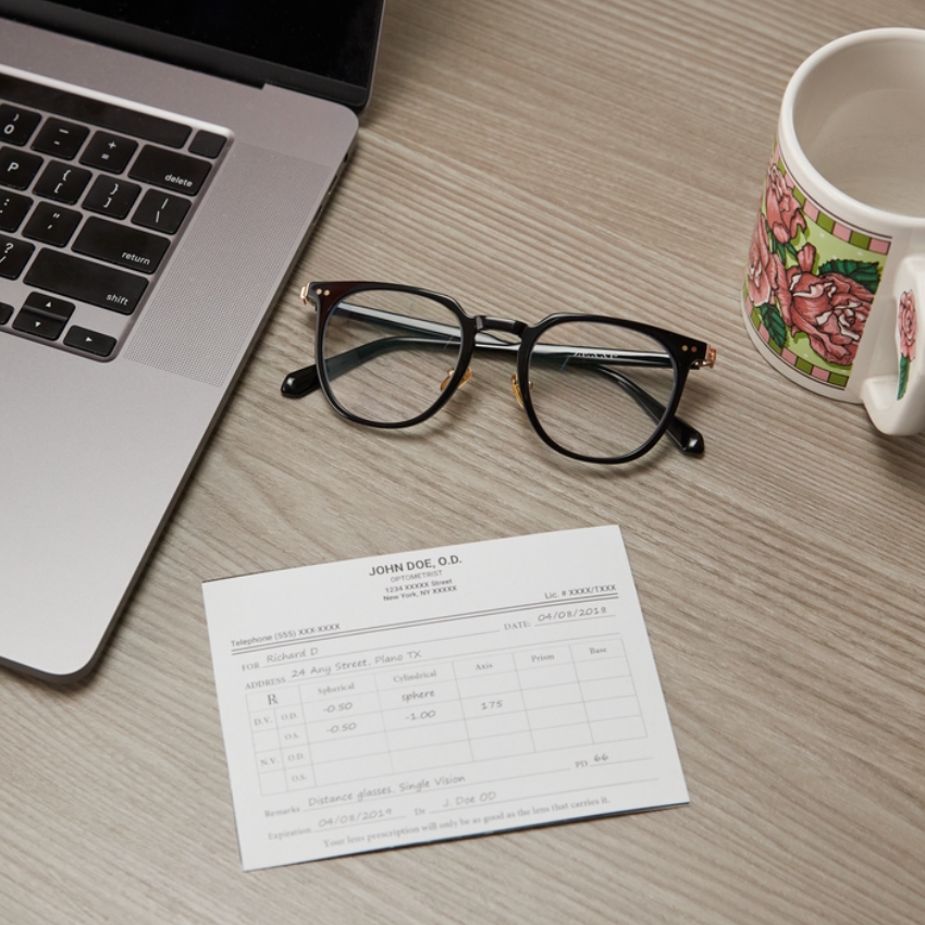 Eyeglasses and optical prescription from John Doe, O.D. on a desk next to a laptop and a floral mug.