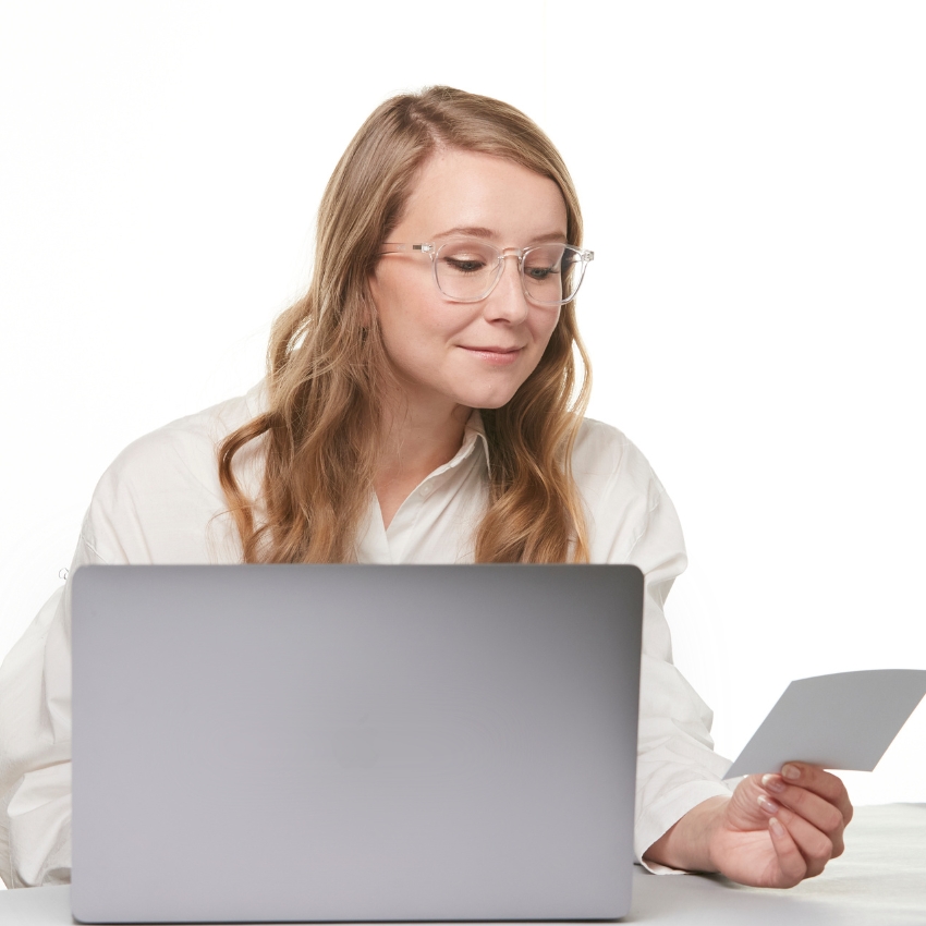 Woman with glasses working on a laptop, holding a card in hand.