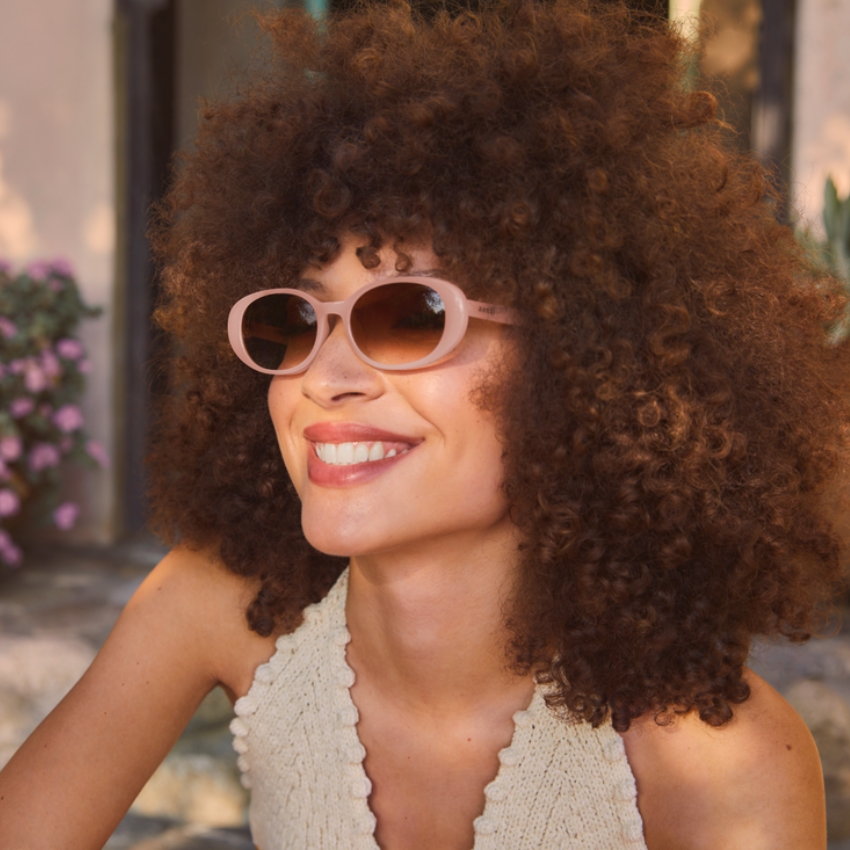 Smiling person wearing pink oval sunglasses with a wavy-edge knit top.