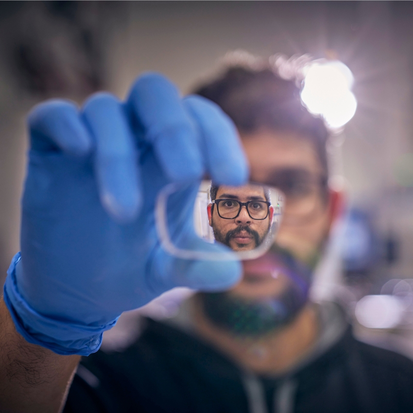 Close-up of a person wearing a blue glove holding a lens. The person's face is reflected in the lens.