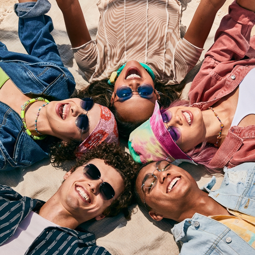 Group of friends wearing colorful sunglasses and bandanas, lying on their backs, smiling and laughing.