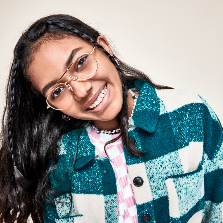 Smiling person wearing gold rimmed glasses, a blue and white checkered jacket, and a beaded necklace.