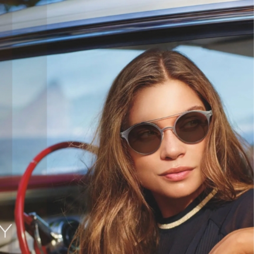 Woman wearing round-frame sunglasses with mirrored lenses, sitting in a car with a red steering wheel.