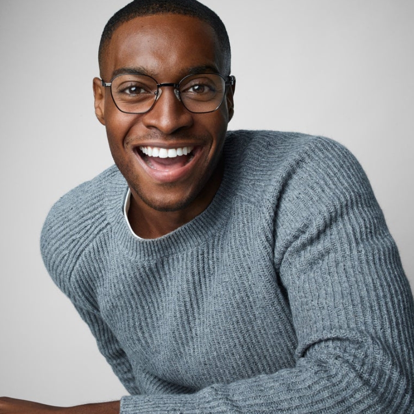 Man wearing black frame glasses and a grey knitted sweater, smiling warmly.