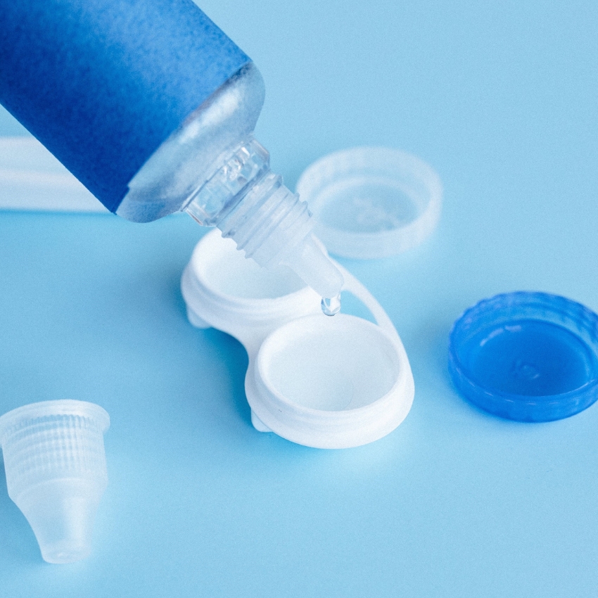 Contact lens solution bottle dispensing drop into an open contact lens case on a blue surface.