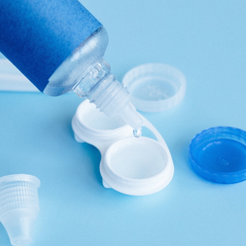 Contact lens solution bottle with a drop falling into a white contact lens case; blue background.