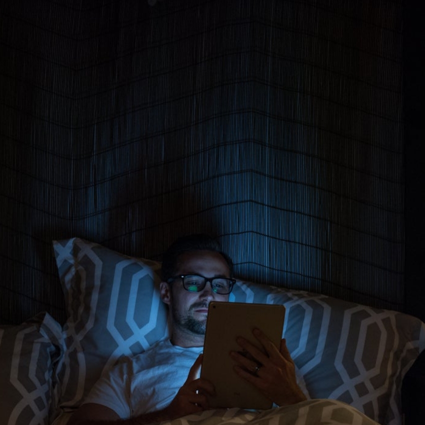 Person using a tablet in bed, illuminated by the screen in a dark room.