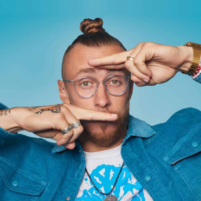 Man with a top knot wearing round glasses, a blue denim jacket over a graphic tee, and posing creatively.