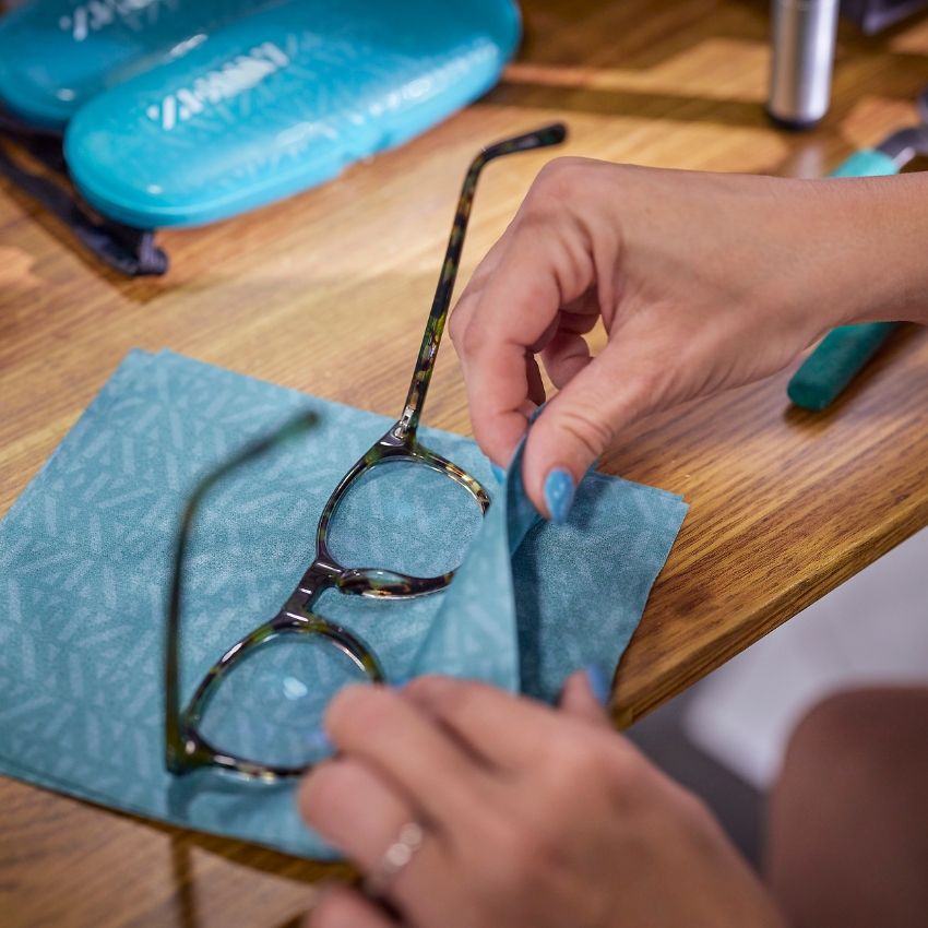 Hands cleaning eyeglasses with a turquoise cloth. Blue cases in the background.