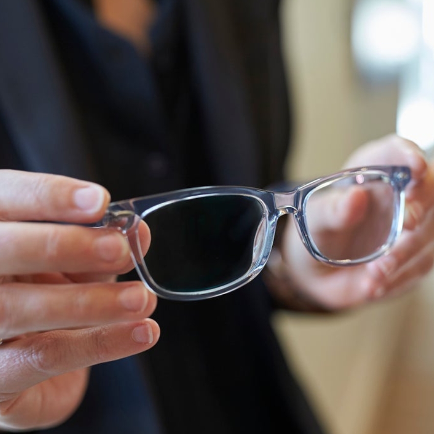 Hands holding a pair of transparent-framed glasses with one dark lens and one clear lens.