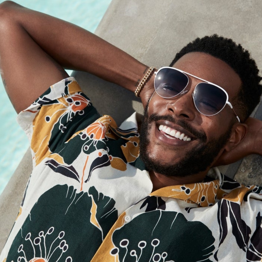 Man wearing floral shirt and oversized aviator sunglasses, smiling while lying down.