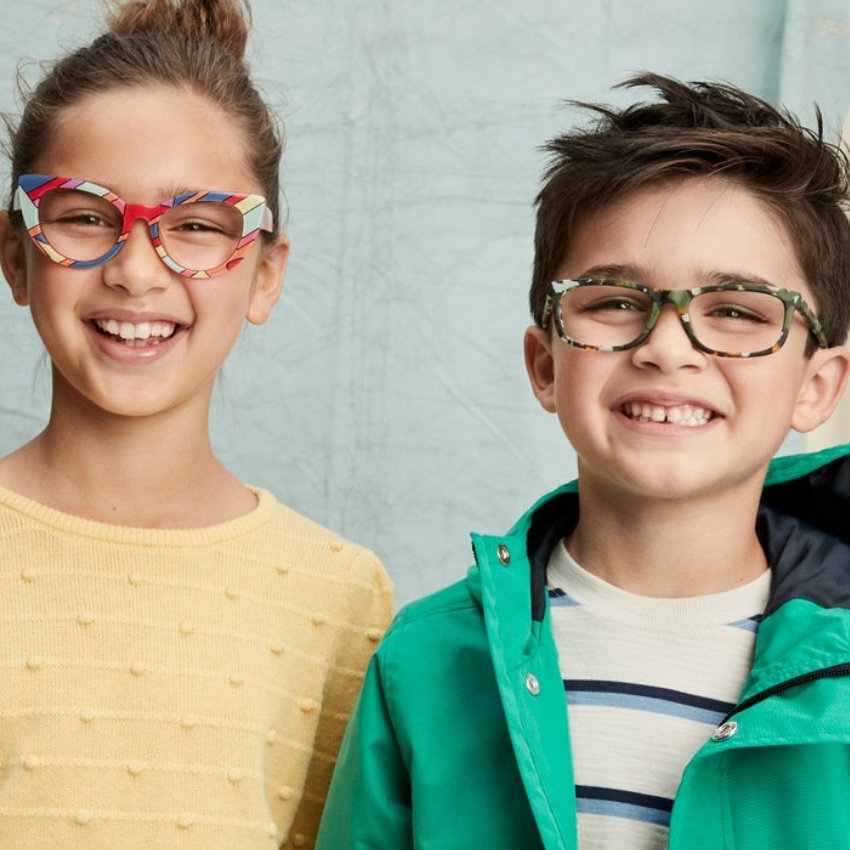 Children wearing colorful, patterned eyeglasses. Girl's glasses are red and multi-colored; boy's glasses are green camo.