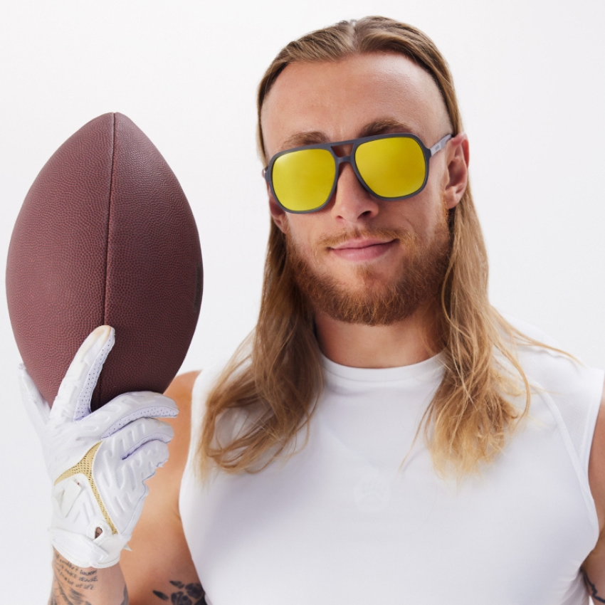 Person with long hair and reflective yellow sunglasses holding a football and wearing a white glove.