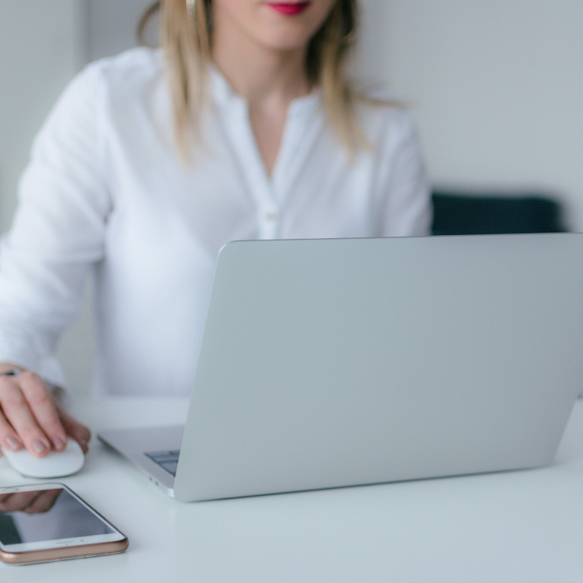 Person using a gray laptop and a white wireless mouse, with a smartphone nearby on a white desk.