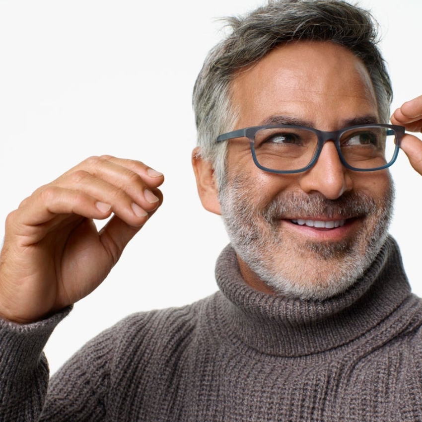 Man adjusting black-frame eyeglasses, smiling, wearing a gray turtleneck sweater.