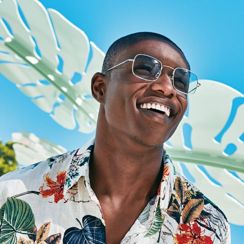 Man wearing square-shaped sunglasses and a floral shirt, smiling under bright sunlight.