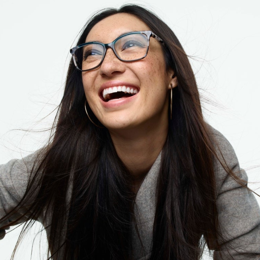 Woman wearing clear-frame eyeglasses and smiling, with long dark hair and large hoop earrings.