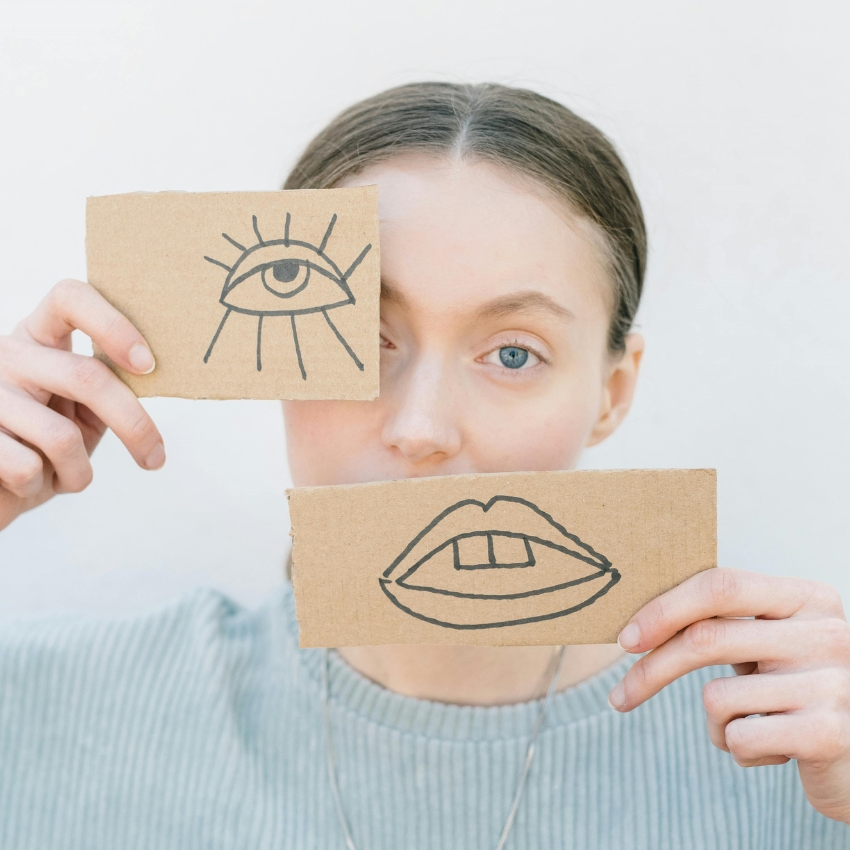 Woman holding two cardboard pieces; one with a drawn eye over her eye and one with drawn lips over her mouth.